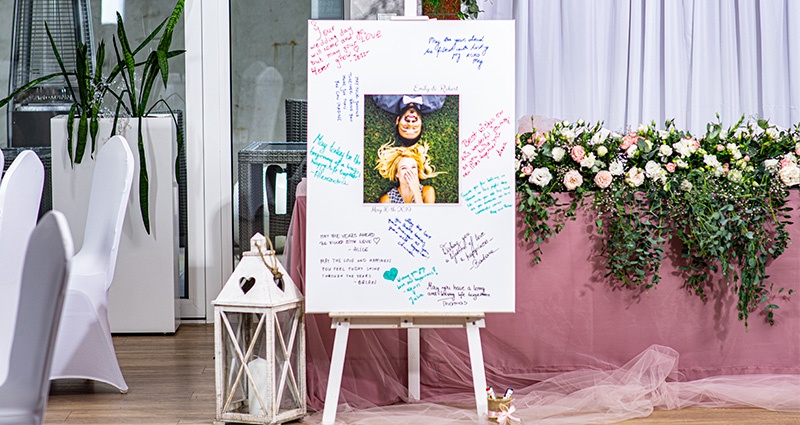 Eine große Leinwand mit Bild eines verliebten Paares in der Mitte, Wünsche der Hochzeitsgäste mit bunten Markern drum herum geschrieben. Daneben eine weiße Laterne und im Hintergrund ein mit pulverrosa Tüll und Blumen geschmückter Tisch