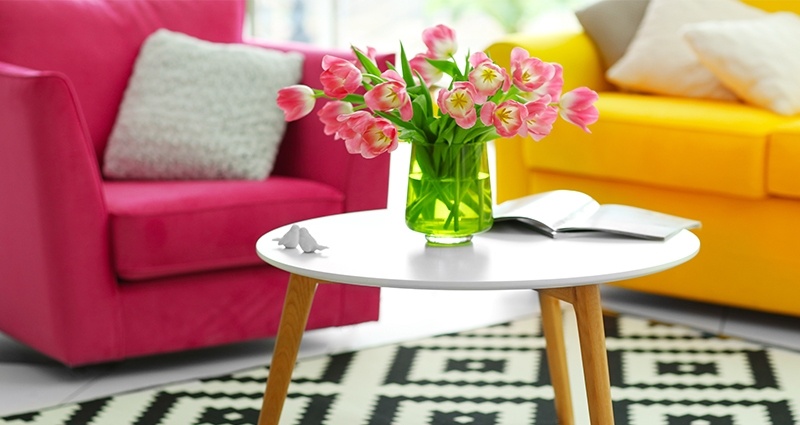 Ein kleiner Tisch im Wohnzimmer, auf dem eine Vase mit rosa Tulpen steht. Darunter liegt ein schwarz- weißer Teppich. Ein aramantfarbener Sessel und ein gelbes Sofa im Hintergrund.